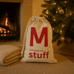 Natural drawstring bag with red text 'M christmas eve stuff' in front of a Christmas tree and fireplace.