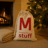 Natural drawstring bag with red text 'M christmas eve stuff' in front of a Christmas tree and fireplace.