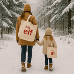Two people walking in a snowy forest with 'big elf' and 'mini elf' tote bags.