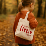 Child wearing a sweater and carrying a tote bag with 'Santa's little helper' text in an autumn setting.