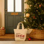 Natural tote bag with 'Santa's Little Helper' text in front of a decorated Christmas tree and door.